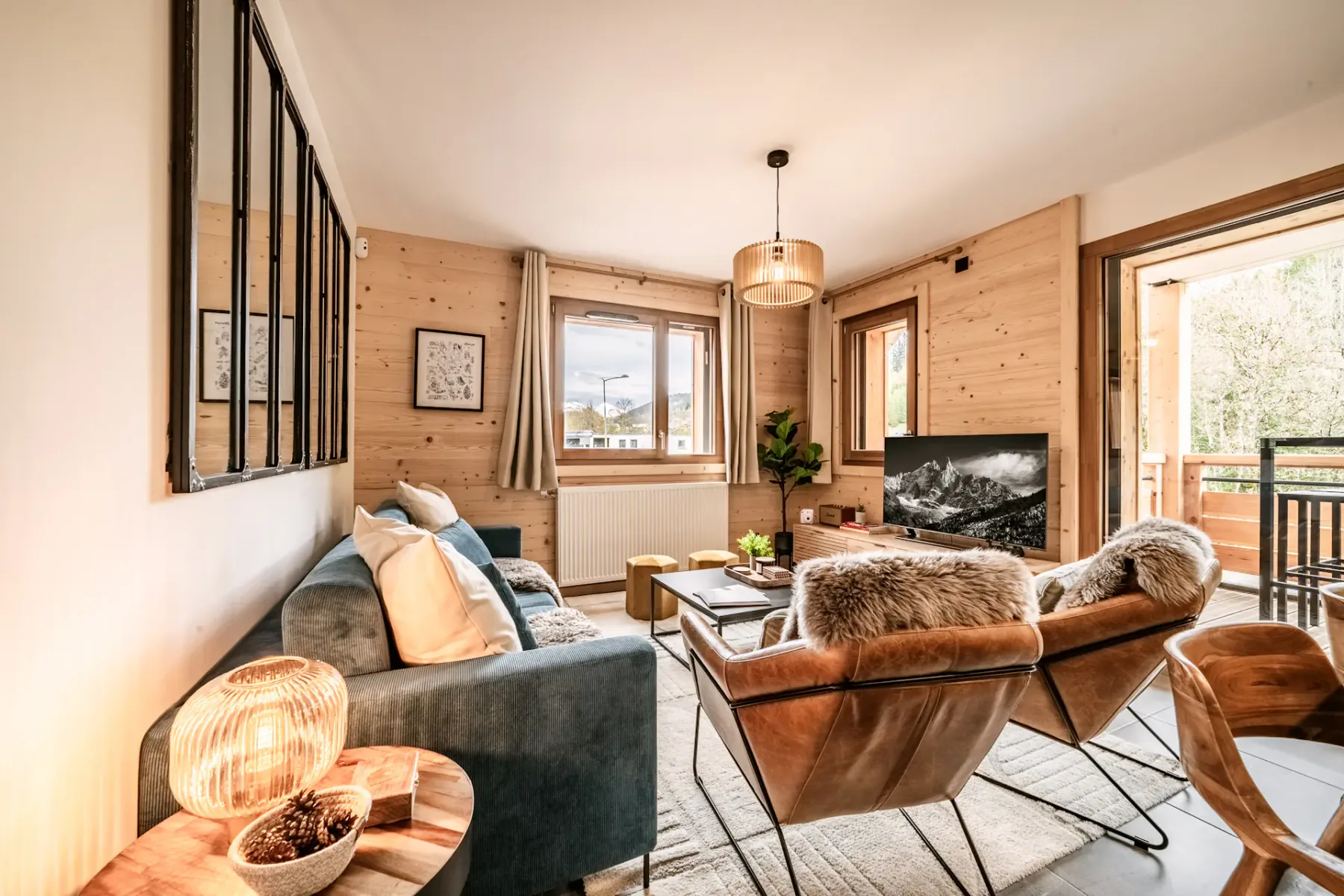 Cozy living room with wooden walls, blue sofa, fur-covered chairs, a small table, and a TV. Large window and balcony door provide natural light.