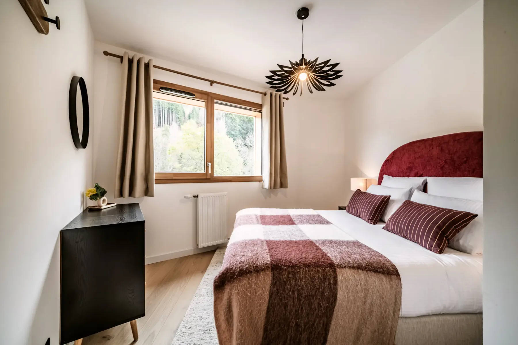 Cozy bedroom with a large bed, plaid blanket, and striped pillows. A unique overhead light fixture and window with curtains brighten the space.