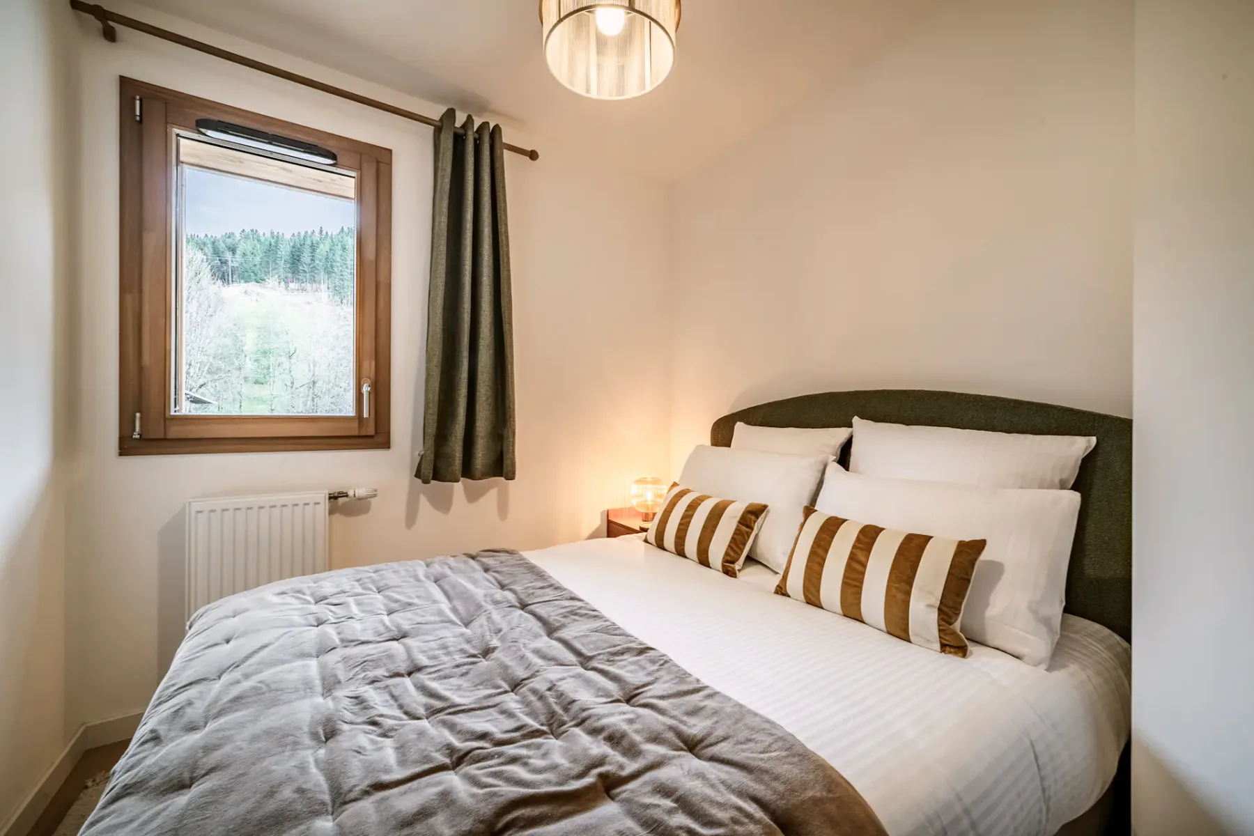 Cozy bedroom with a bed, striped pillows, wooden dresser, round mirror, pendant light, and a window with green curtains.