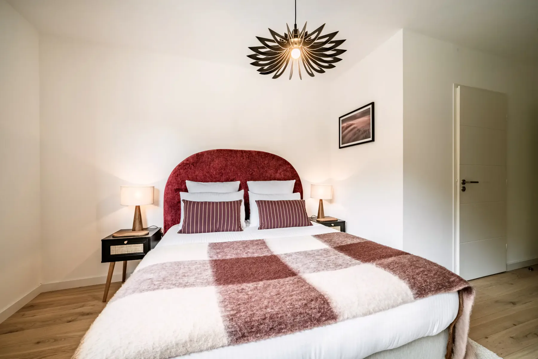 Cozy bedroom with a large bed, plaid blanket, and striped pillows. A unique overhead light fixture and window with curtains brighten the space.