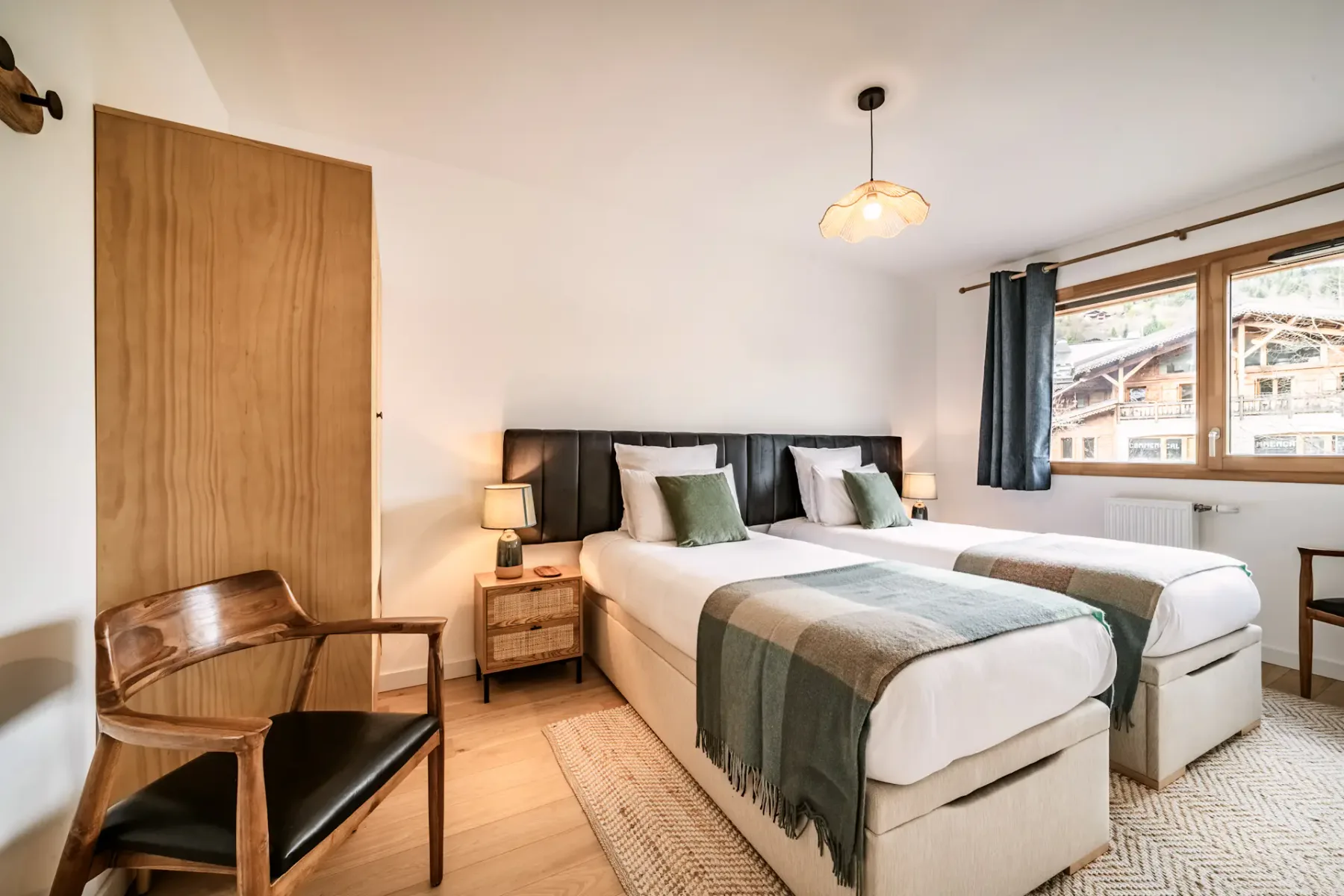 Bright bedroom with two twin beds, green and white bedding, wooden chair, nightstand with lamps, and a window with a view of buildings outside.