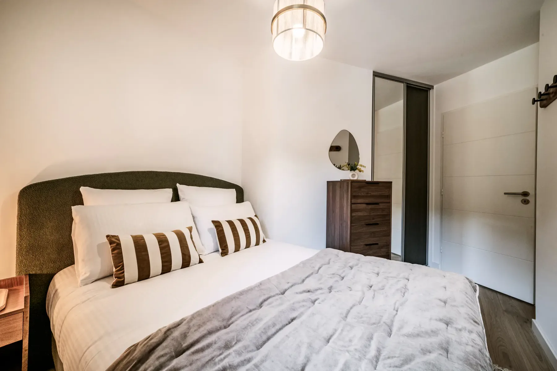 Cozy bedroom with a bed, striped pillows, wooden dresser, round mirror, pendant light, and a window with green curtains.