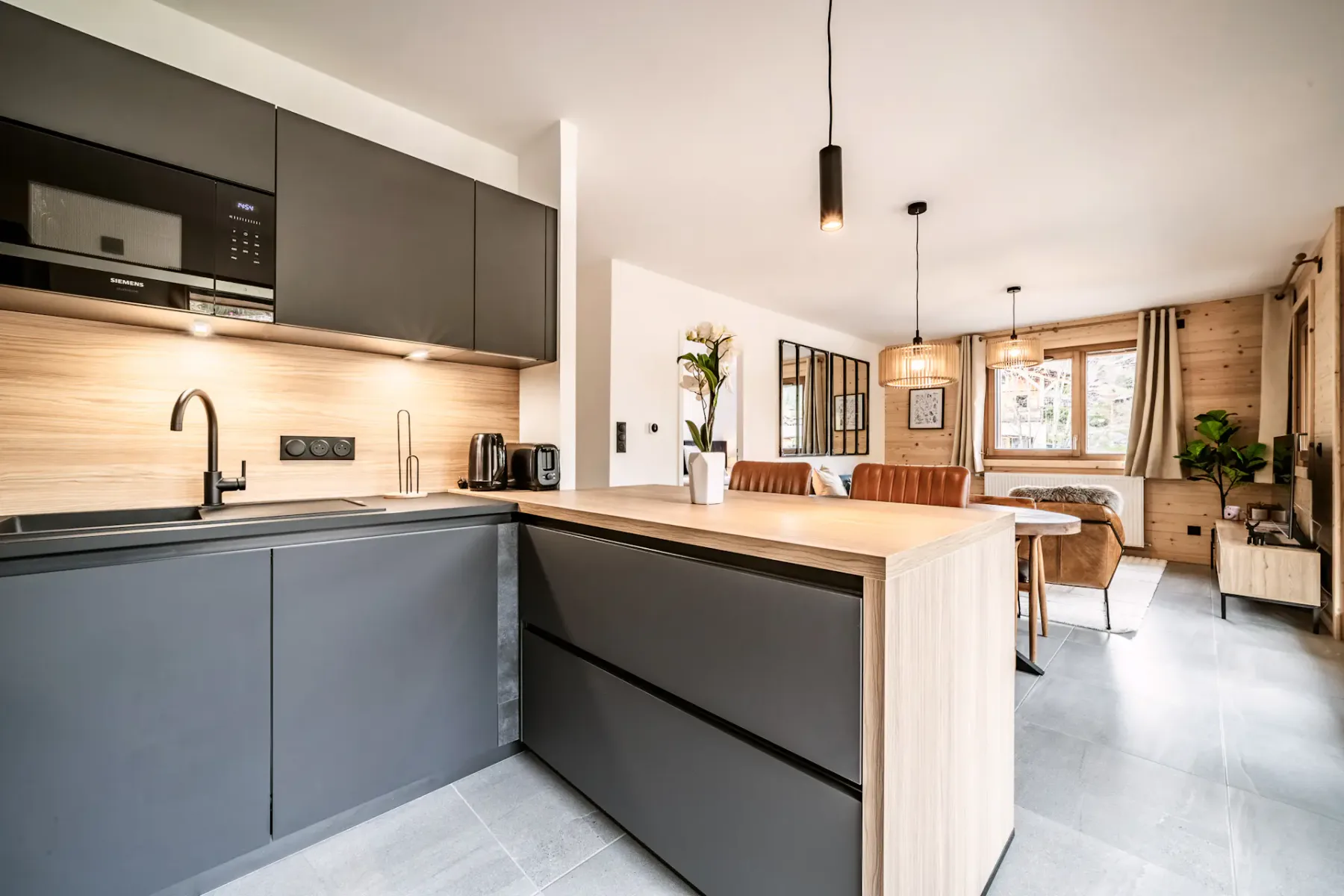Modern kitchen with dark cabinets, wooden island with two brown stools, pendant light, and wall clock. Bright and spacious with light flooring.