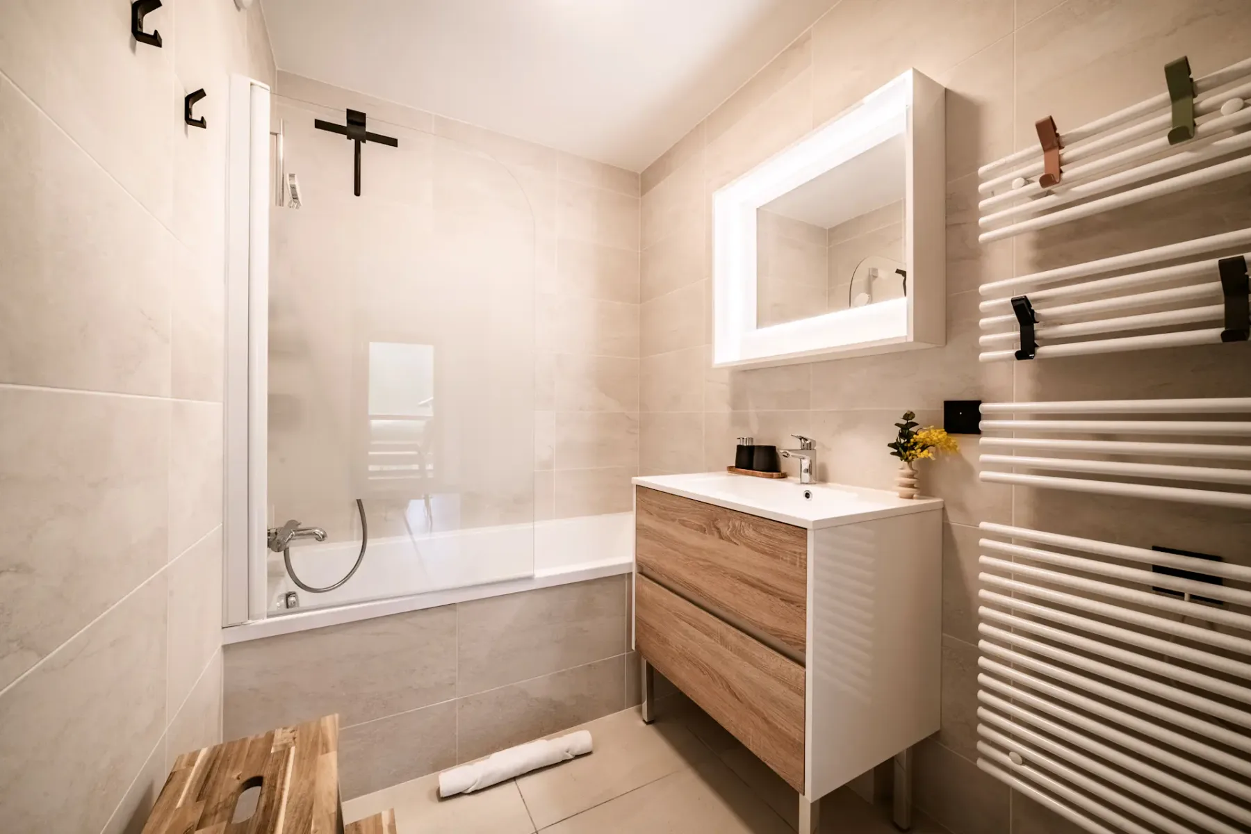 Modern bathroom with a wooden vanity, illuminated mirror, towel radiator, bathtub with glass screen, and beige tile walls.