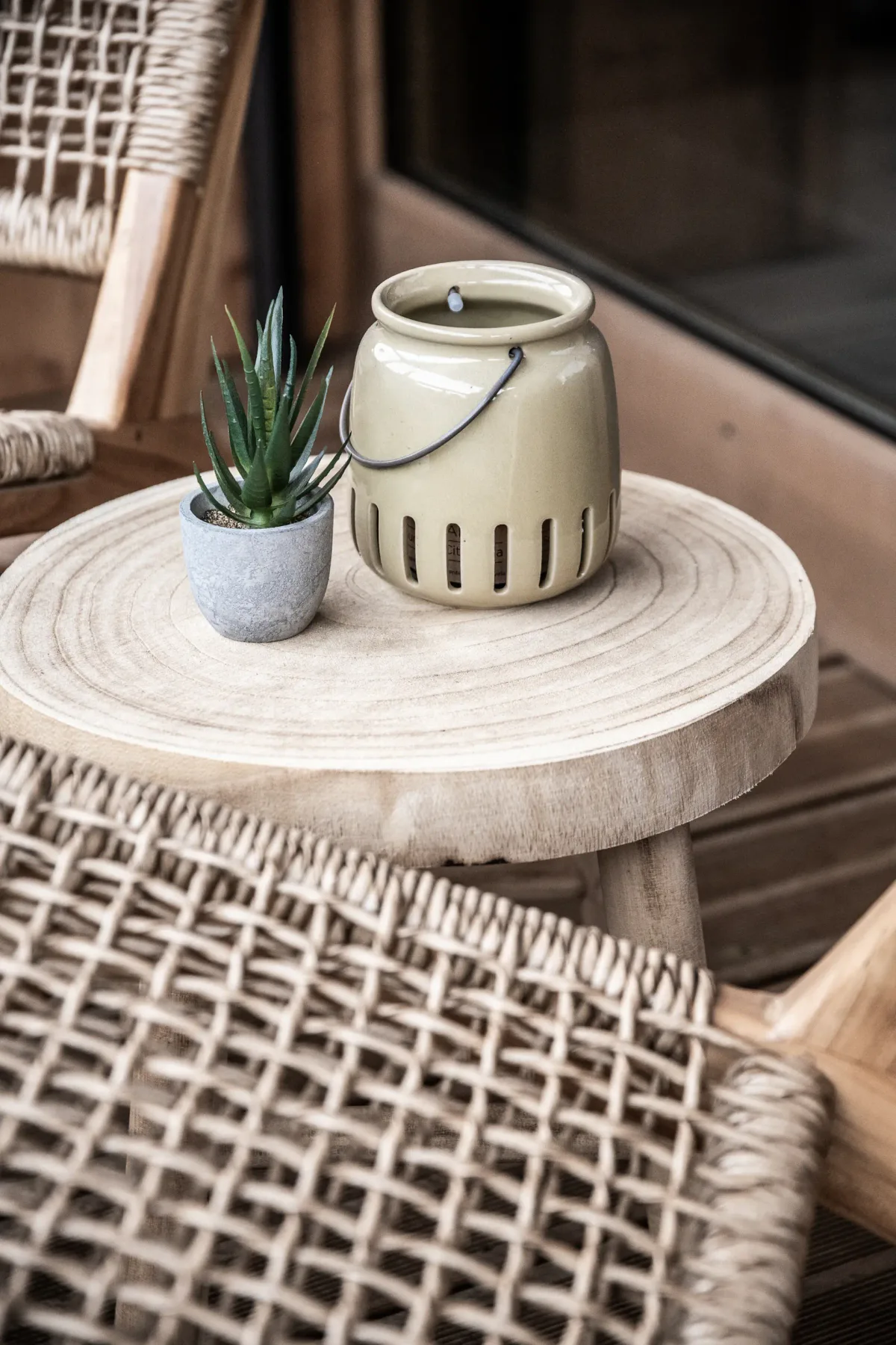Woven chairs surround a wooden table with a small potted succulent and a beige lantern, creating a cozy outdoor setting.