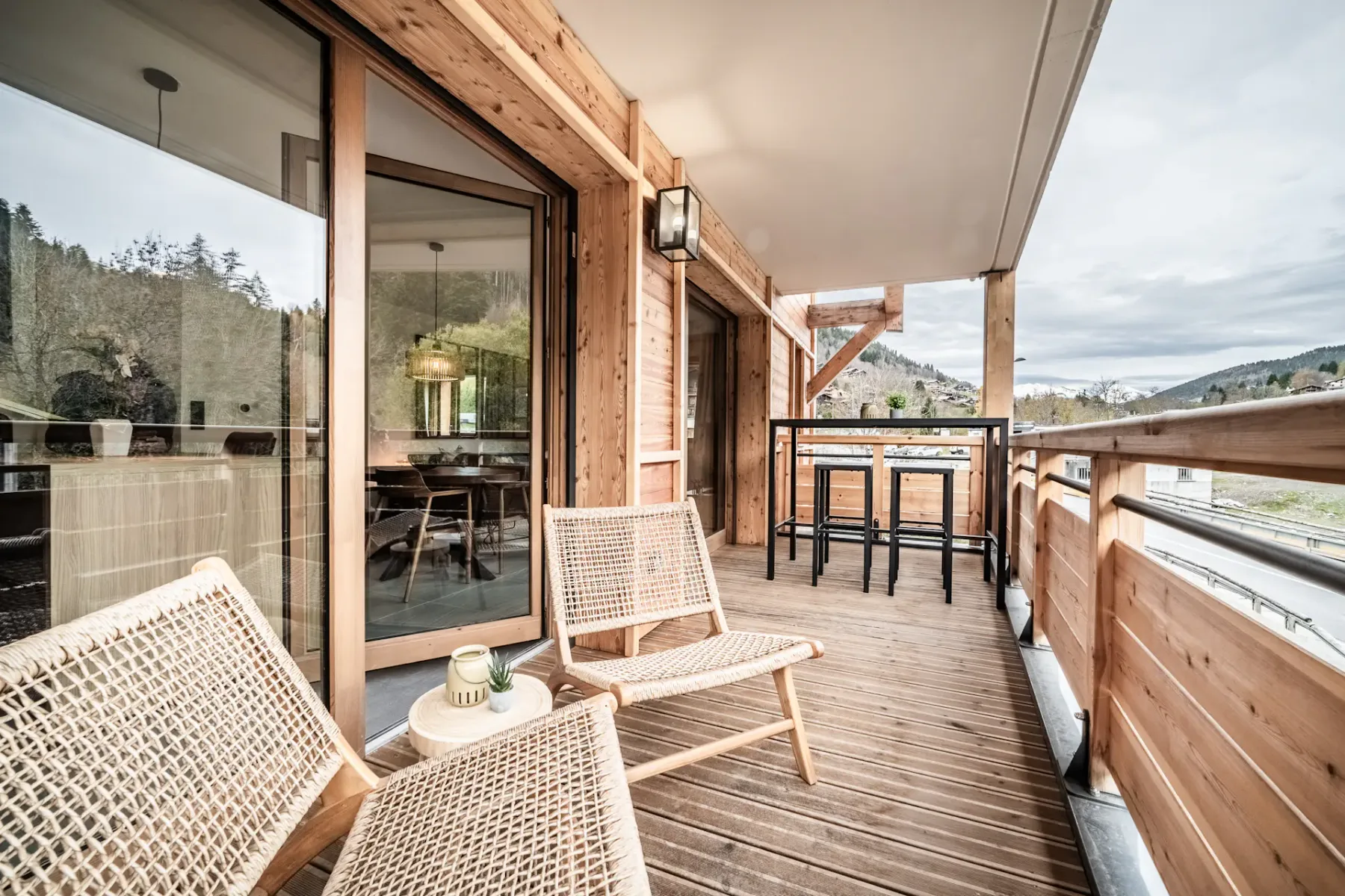 Wooden balcony with a tall black table and stools, overlooking a lush forest landscape under a cloudy sky.