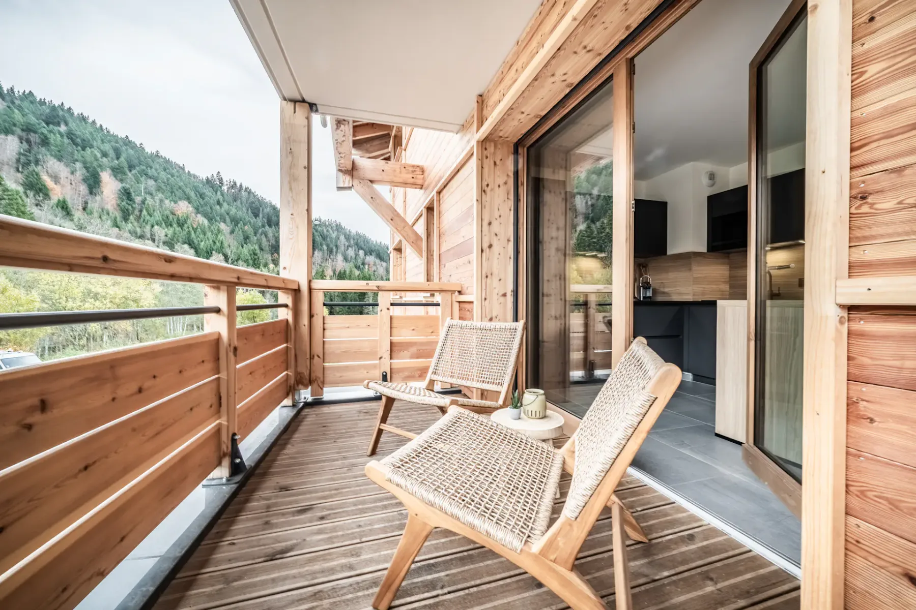 Wooden balcony with a tall black table and stools, overlooking a lush forest landscape under a cloudy sky.