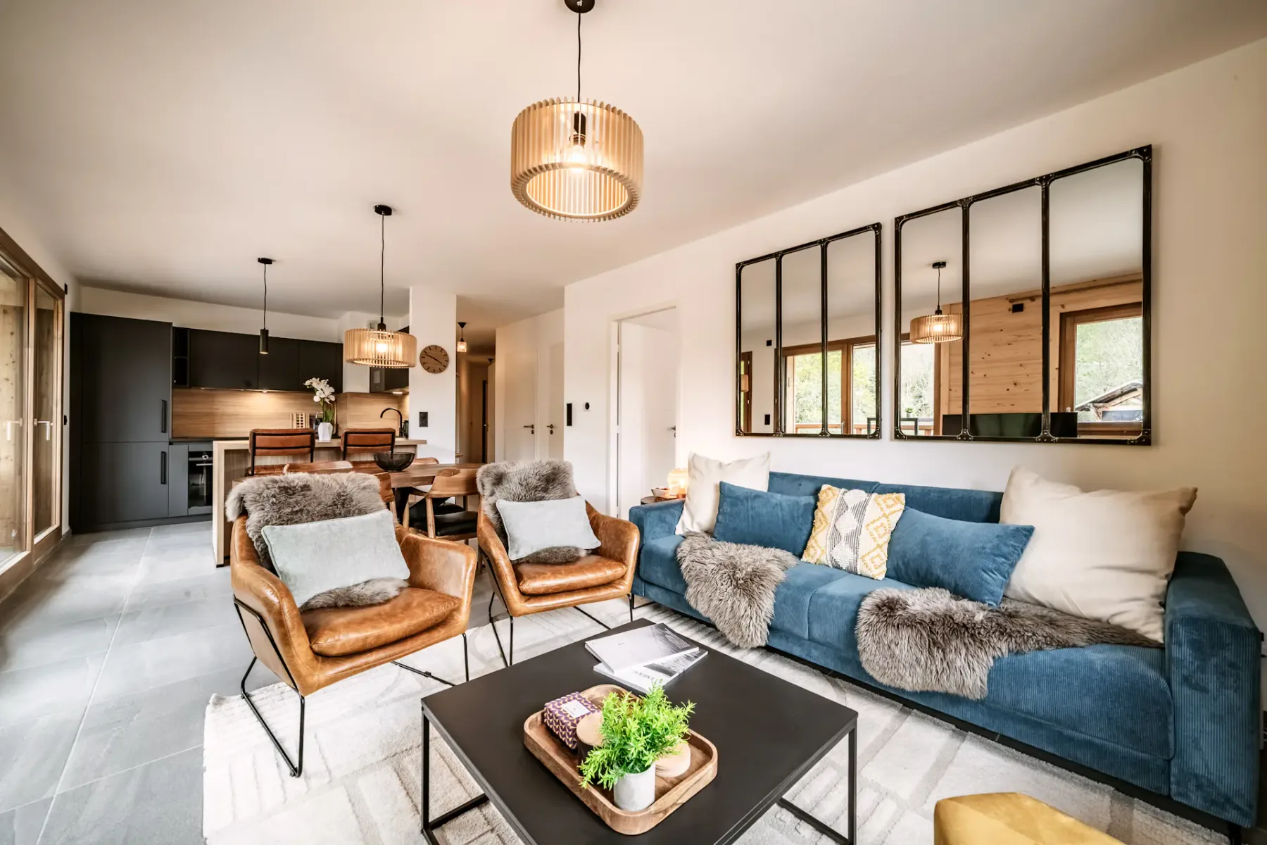 Cozy living room with wooden walls, blue sofa, fur-covered chairs, a small table, and a TV. Large window and balcony door provide natural light.
