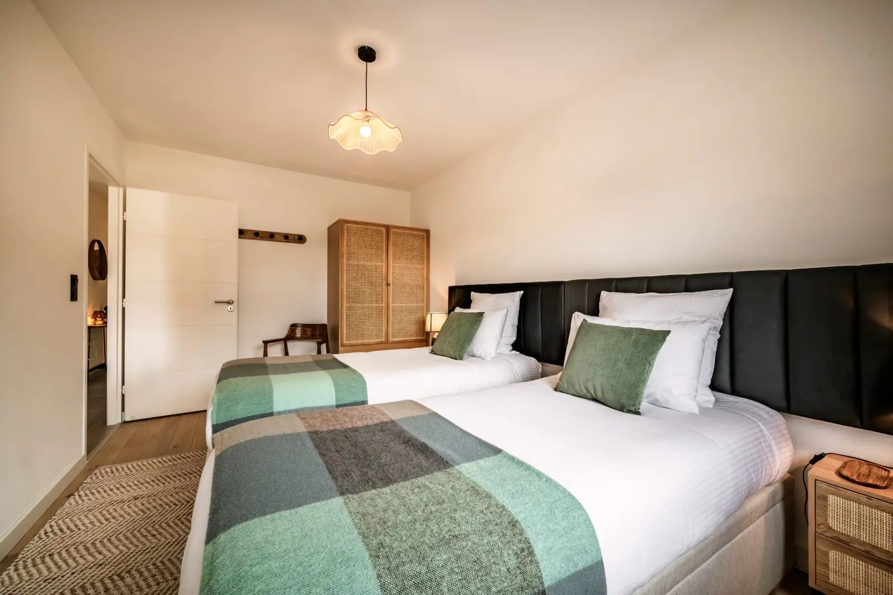 Bright bedroom with two twin beds, green and white bedding, wooden chair, nightstand with lamps, and a window with a view of buildings outside.