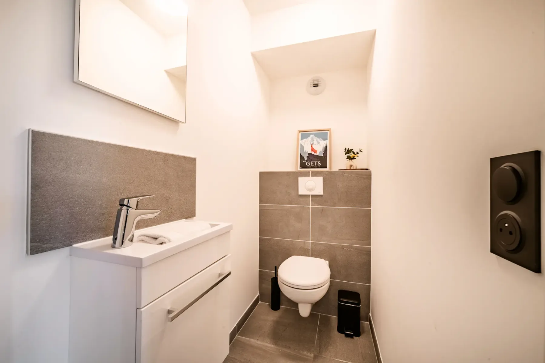 Modern bathroom with a wall-mounted toilet, small sink with a faucet, mirror, and framed artwork. Neutral tones and minimalist design.