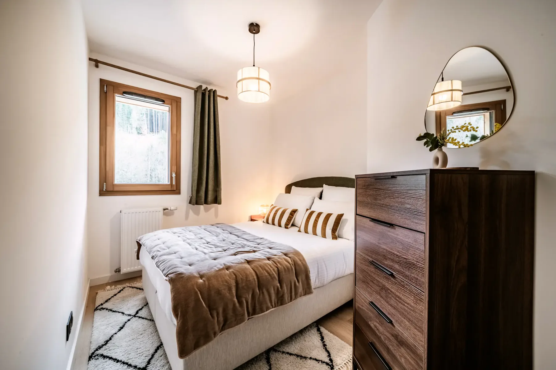 Cozy bedroom with a bed, striped pillows, wooden dresser, round mirror, pendant light, and a window with green curtains.
