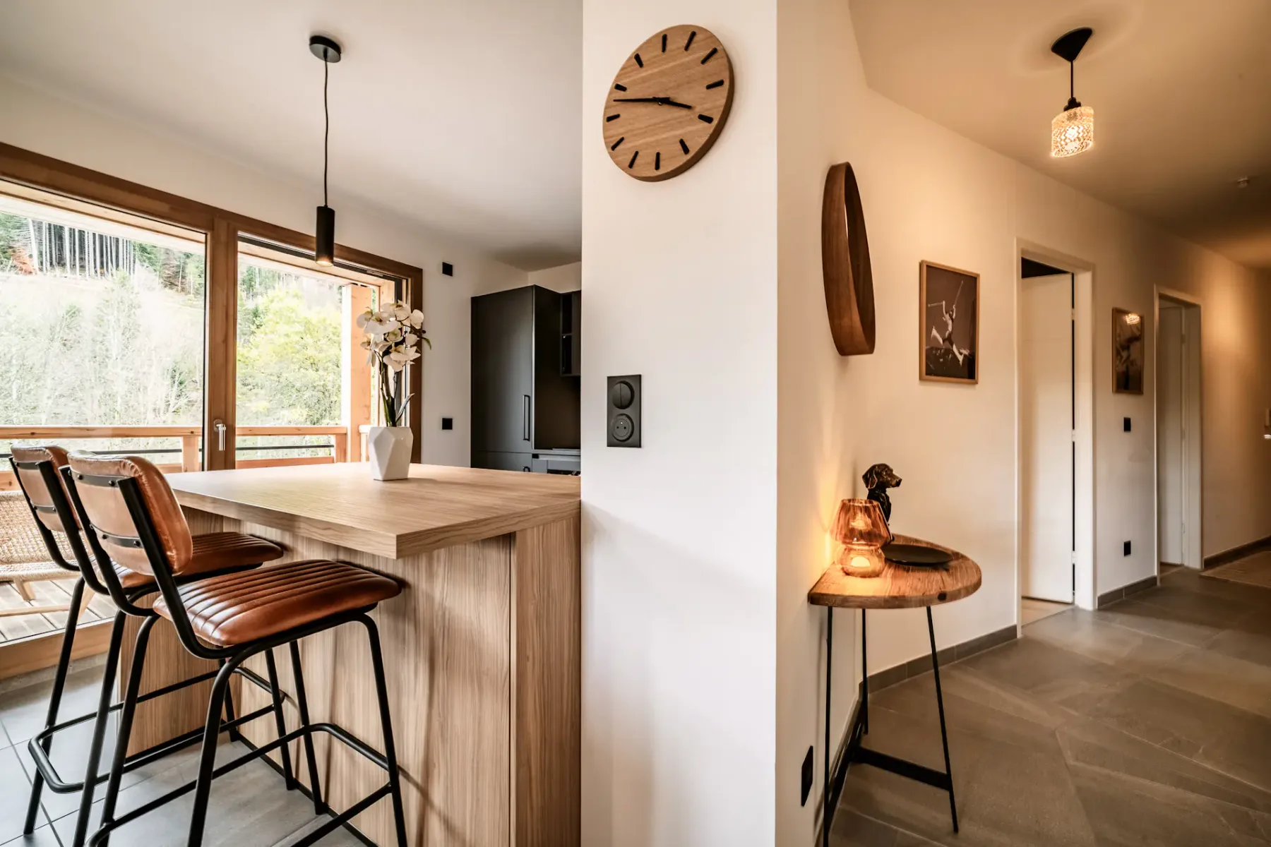 Modern dining area with a round wooden table, leather chairs, and a kitchen island. Large windows overlook a balcony with greenery outside.