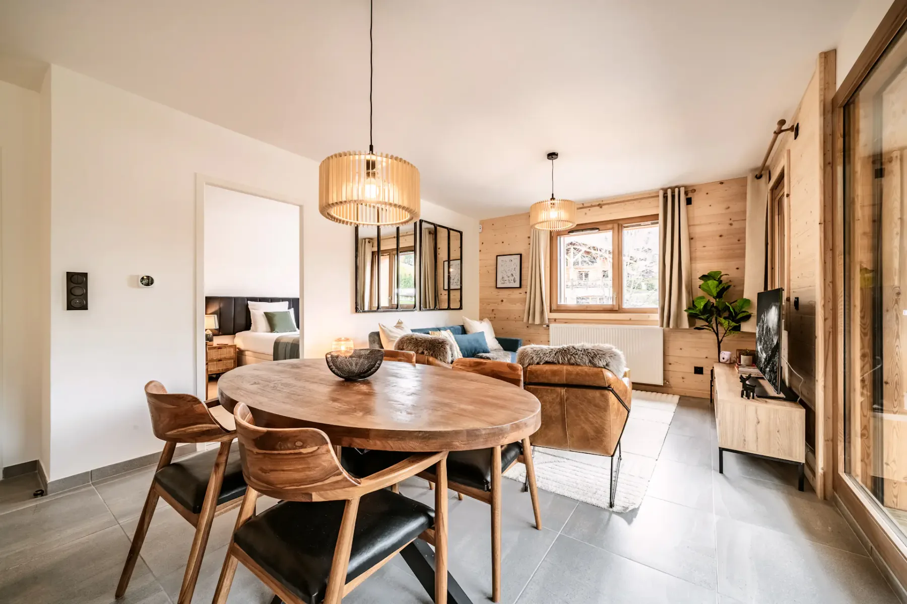 Modern dining area with a round wooden table, leather chairs, and a kitchen island. Large windows overlook a balcony with greenery outside.