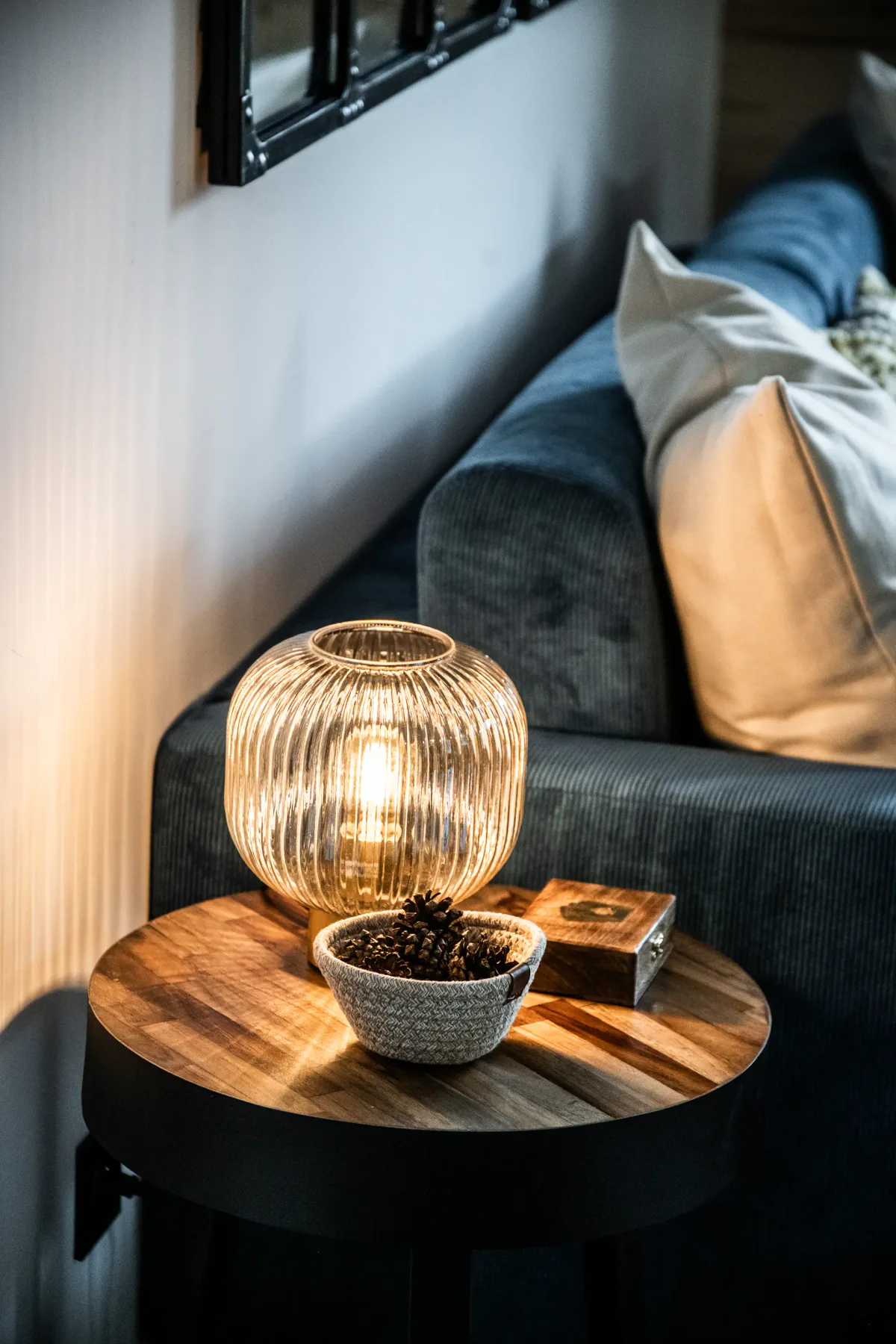 A cozy living room corner with a lit ribbed glass lamp, a small wooden table, a bowl of pinecones, and a blue sofa with white cushions.