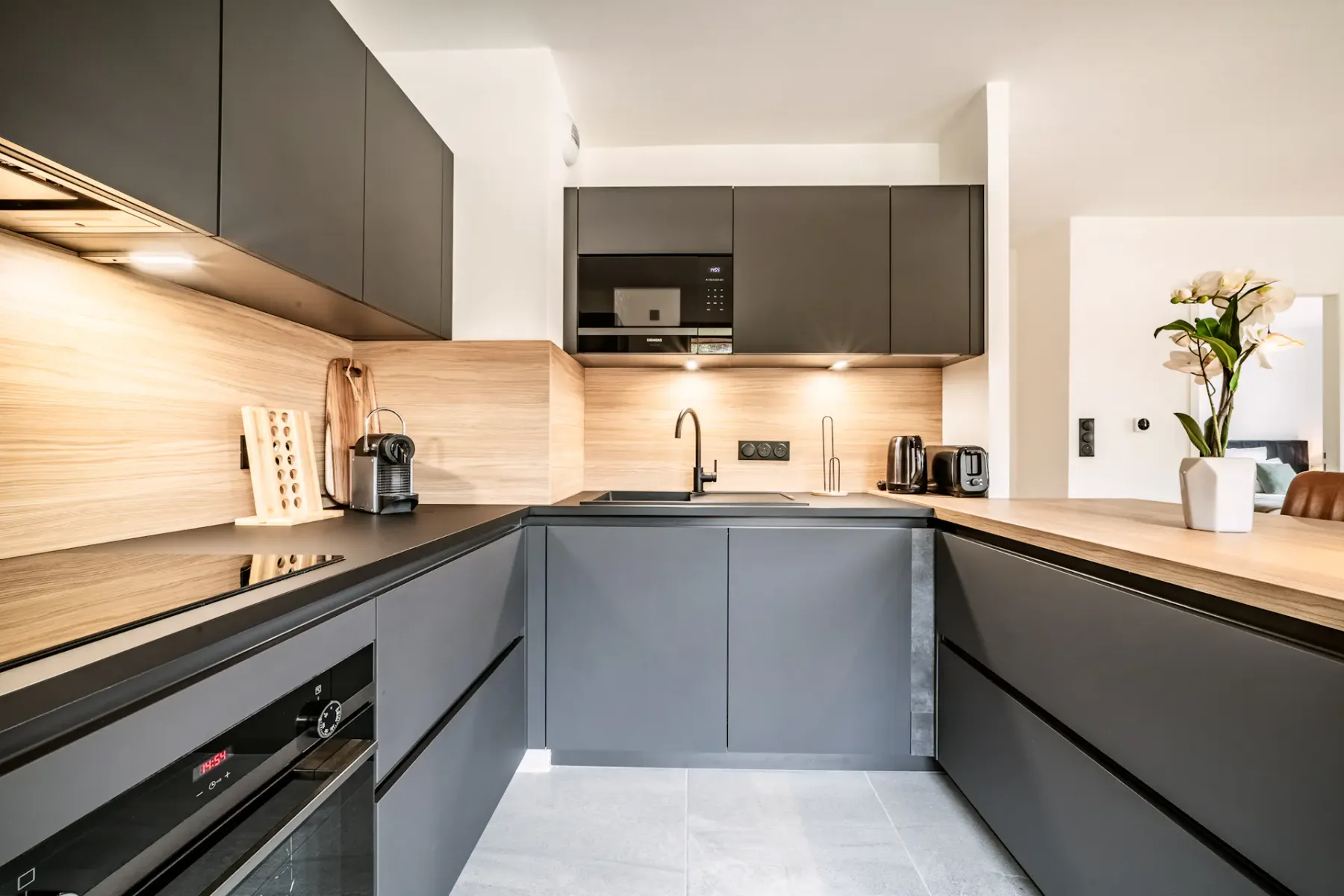 Modern kitchen with dark cabinets, wooden island with two brown stools, pendant light, and wall clock. Bright and spacious with light flooring.