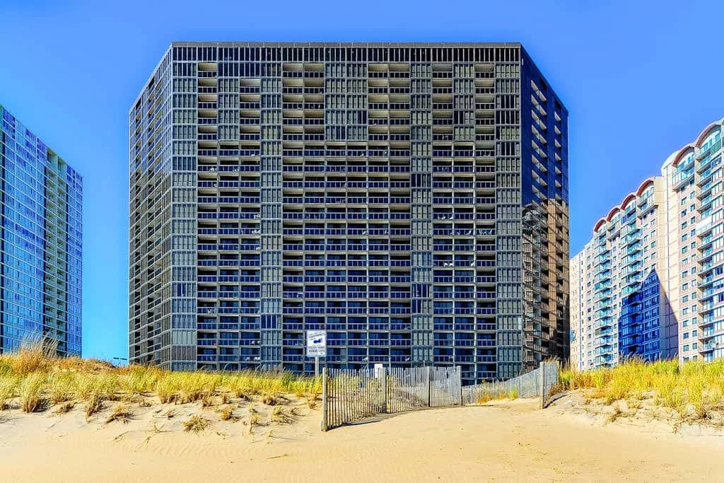 A tall apartment building stands behind sandy dunes under a clear blue sky.