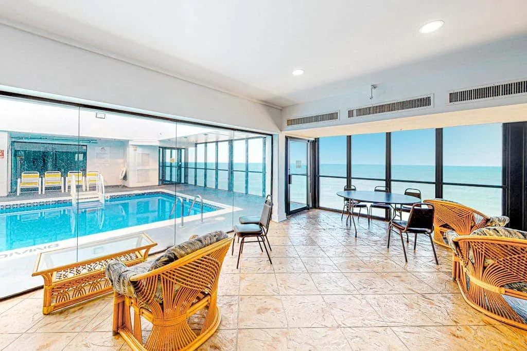 Indoor poolside lounge with wicker seating and large windows overlooking the water.