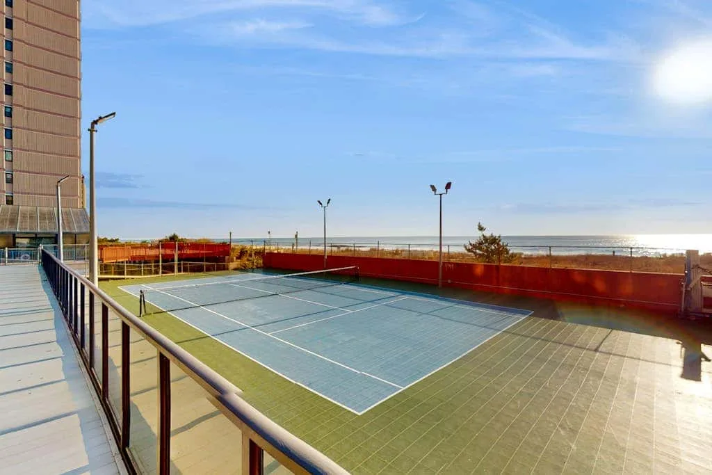 An outdoor tennis court sits beside a building, with the sea visible beyond it.