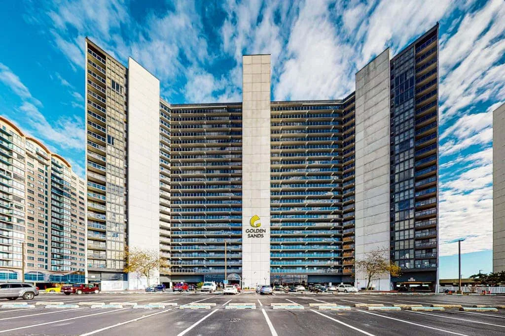 A high-rise building faces a paved parking area beneath a bright, cloud-filled sky.