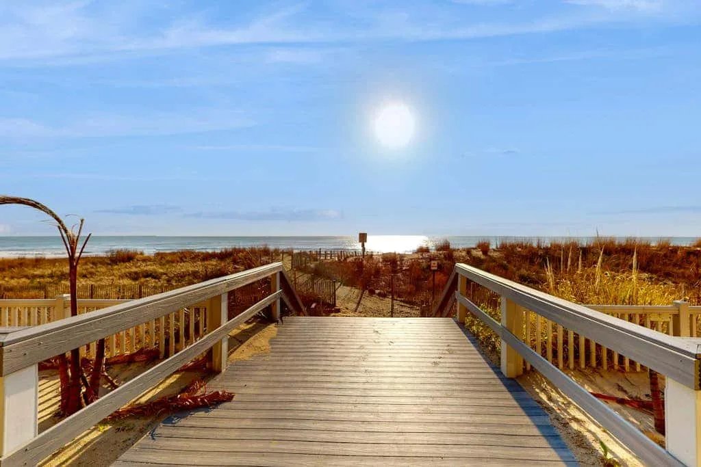 A wooden boardwalk crosses beach grass toward the ocean under a bright sky.
