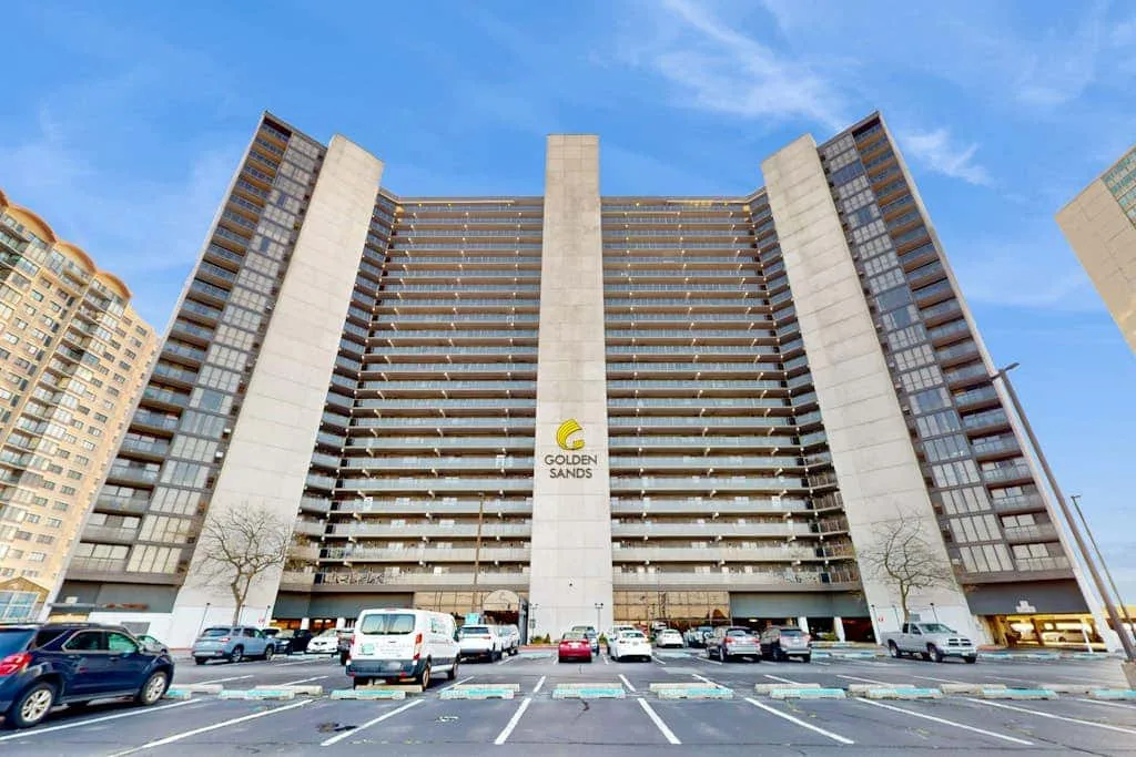 A tall apartment building rises above a parking lot beneath a blue sky.