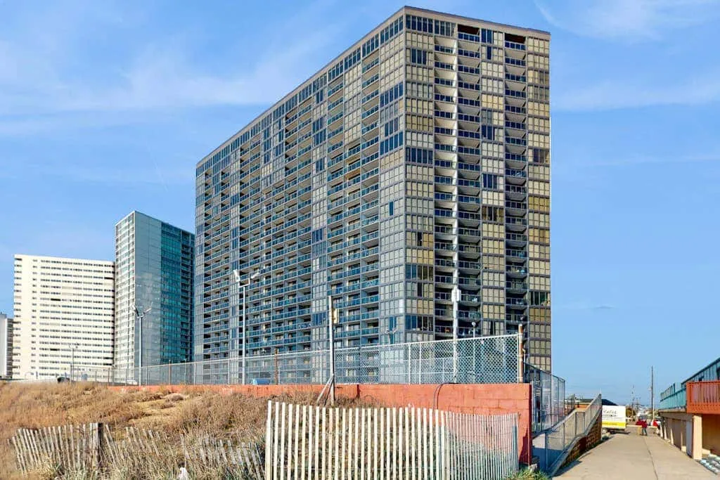 A tall apartment building stands beside a fenced walkway under a clear sky.