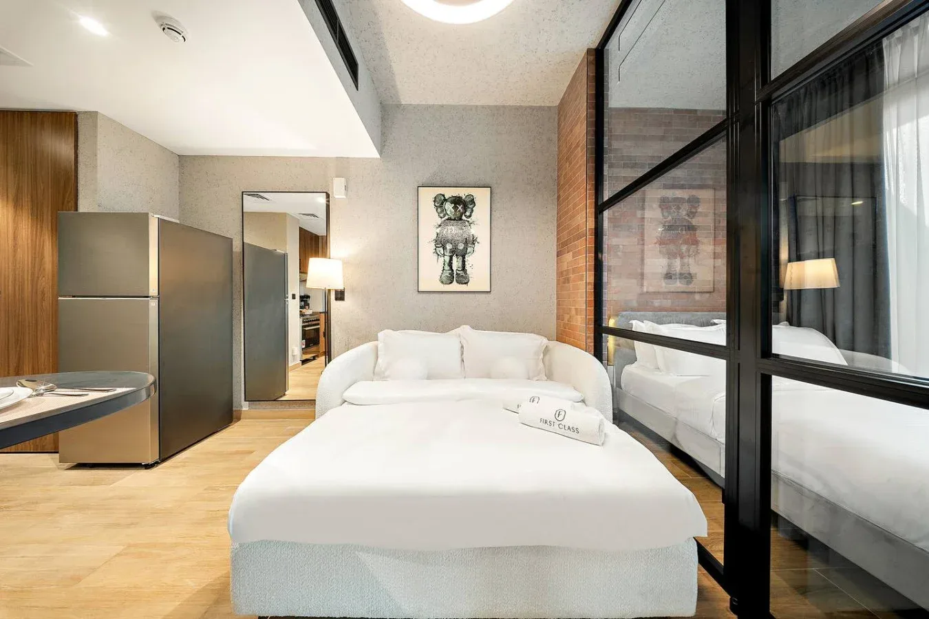 Un dormitorio luminoso con una cama blanca, una pequeña cocina y un separador de vidrio.