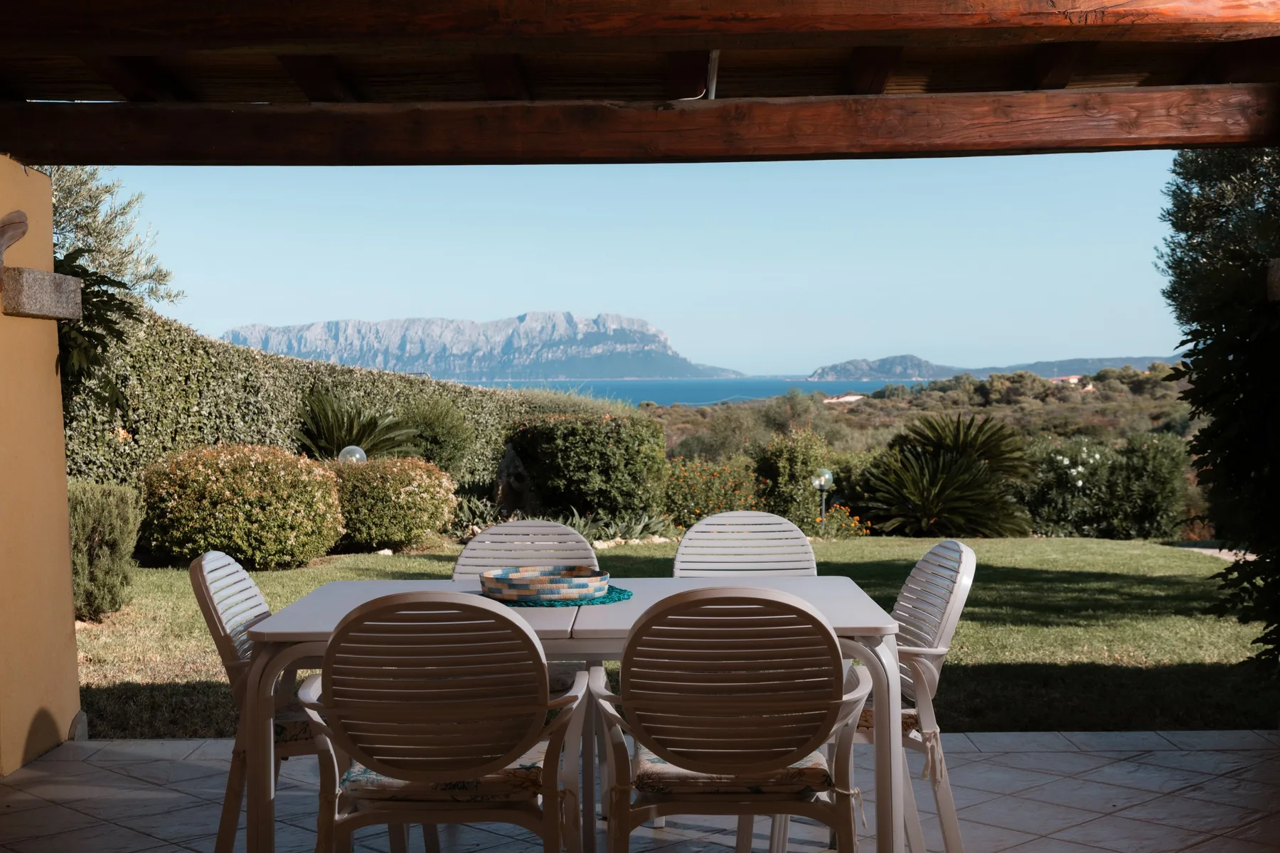 Una zona de comedor al aire libre bajo una pérgola de madera mira a un jardín y agua a lo lejos.