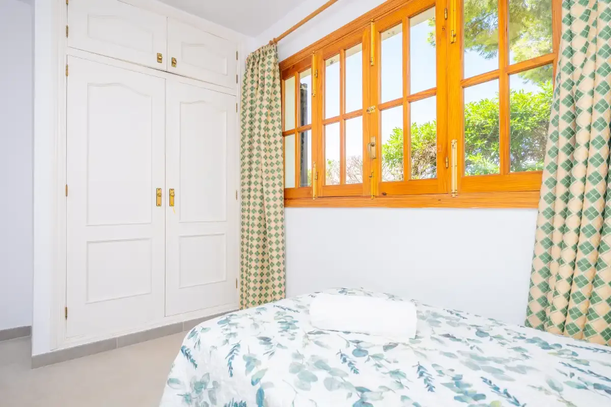 A bright bedroom with a bed beneath wooden-framed windows and patterned curtains.