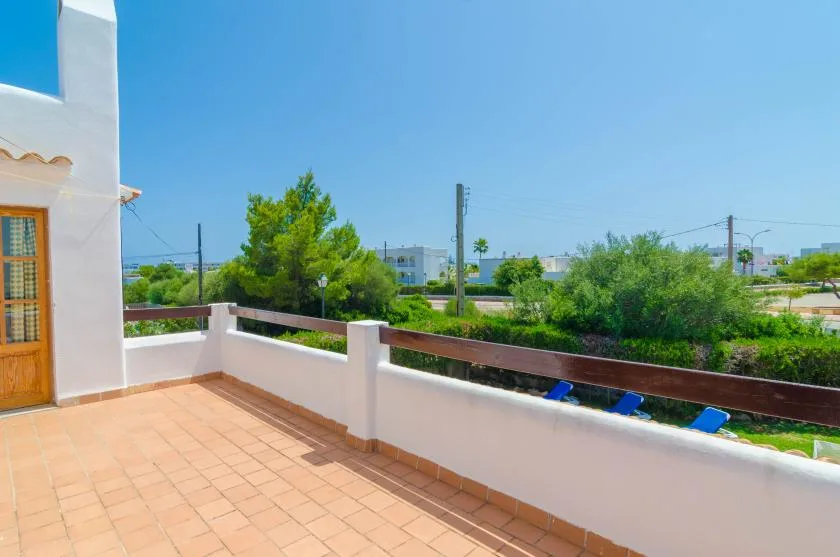 A tiled outdoor terrace overlooks green trees and low buildings beneath a clear blue sky.