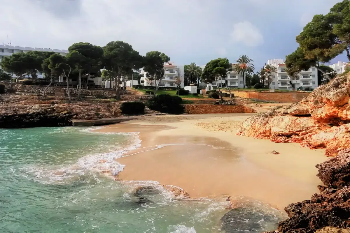 A sandy cove with turquoise water, rocky edges, and buildings among trees.