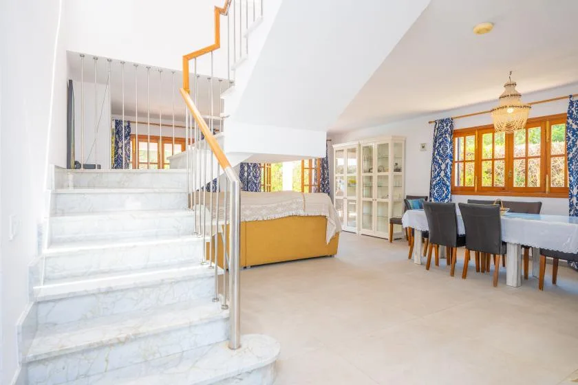 A bright open room with a staircase, seating, and a dining table by the windows.