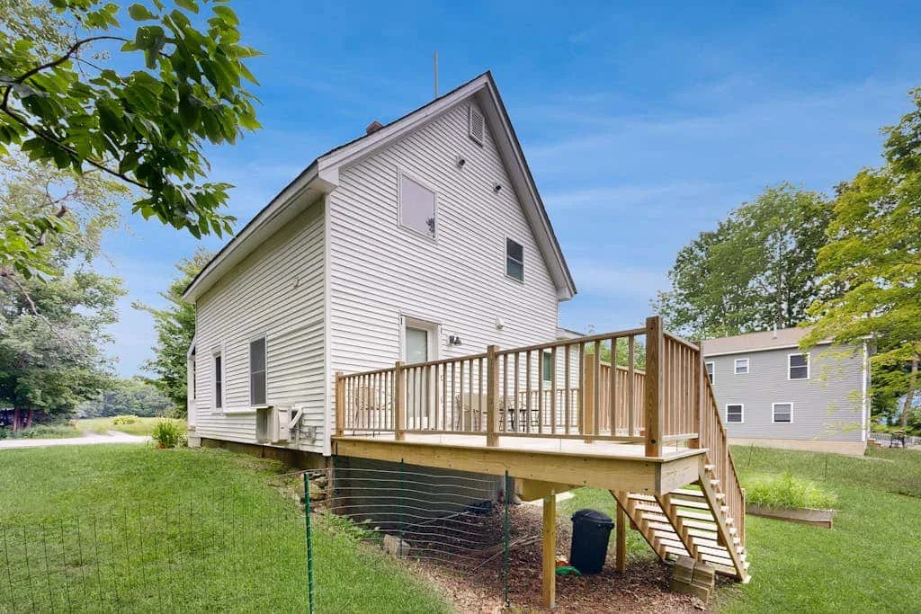 A white two-story house with a wooden deck and stairs in a grassy yard.