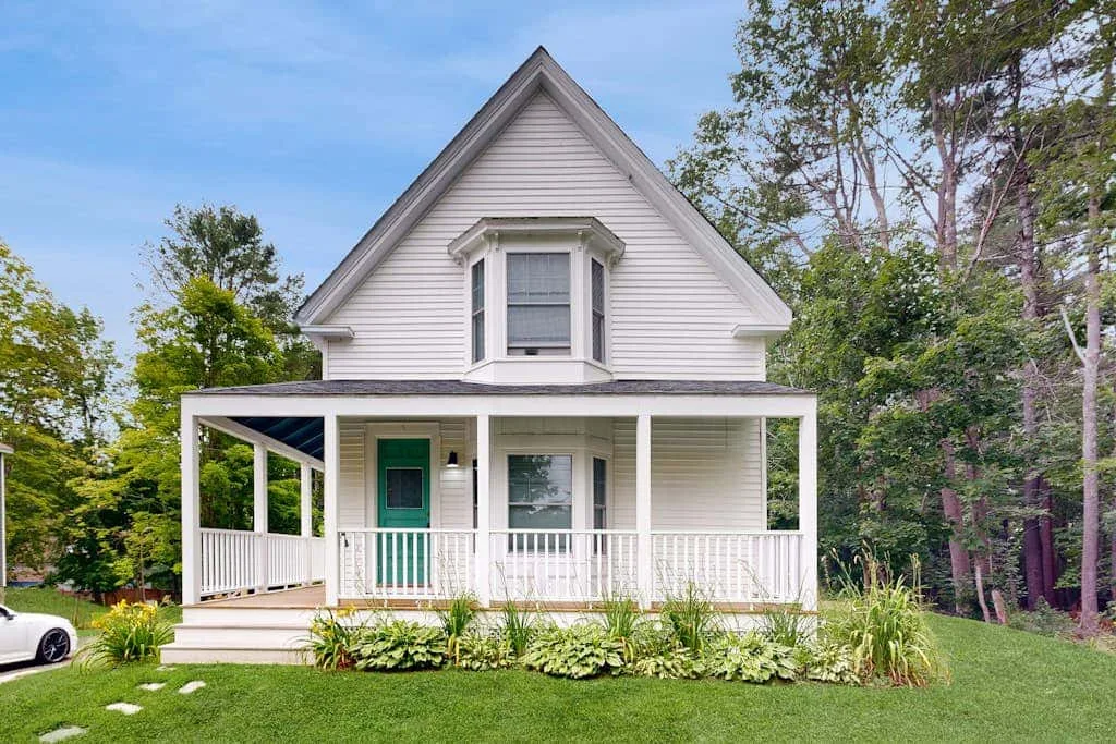 A white house with a covered porch stands among trees and a green lawn.