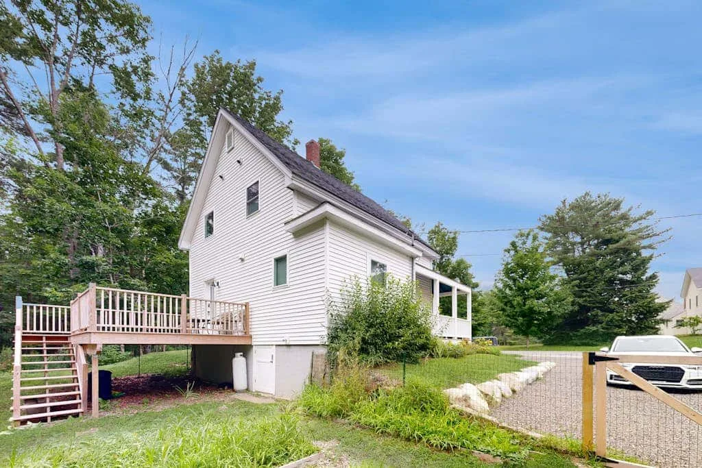 A white house stands beside a gravel driveway, with a wooden deck and surrounding trees.