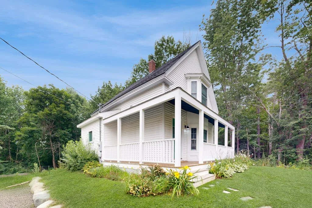 A white house with a covered porch stands on a grassy lawn among trees.