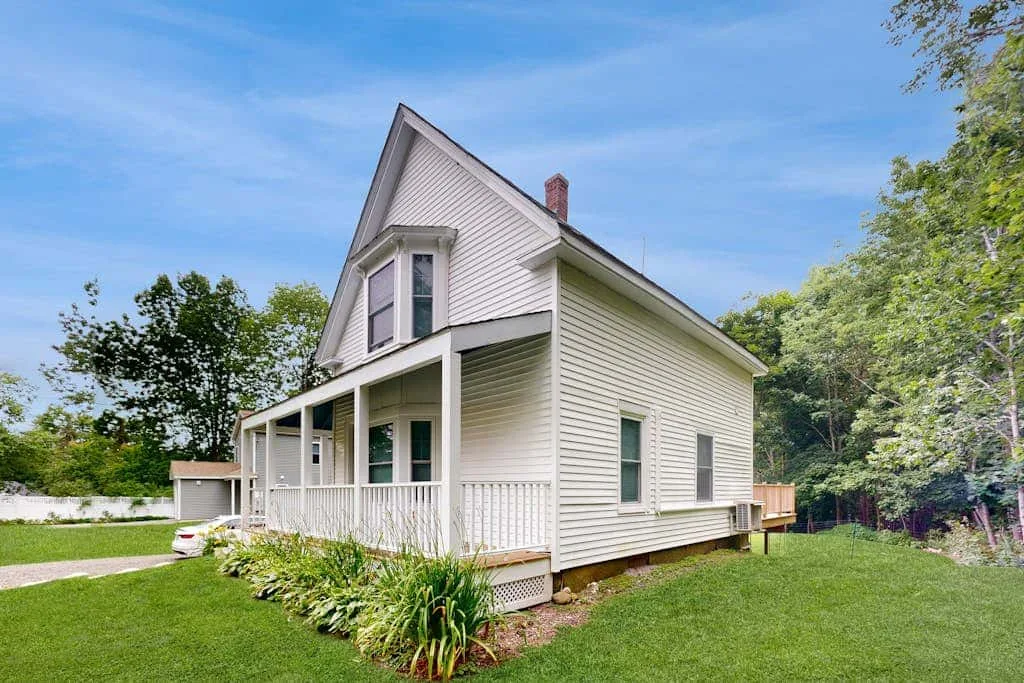 A light-colored house with a covered porch sits beside a green lawn.