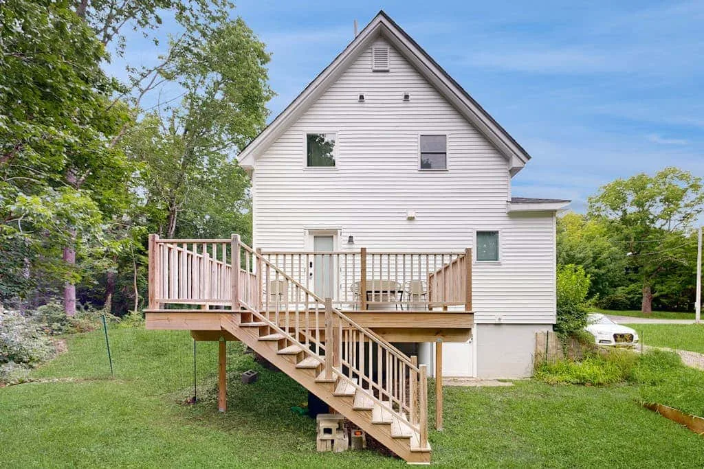 A white house with a raised wooden deck and stairs opens onto a grassy yard.
