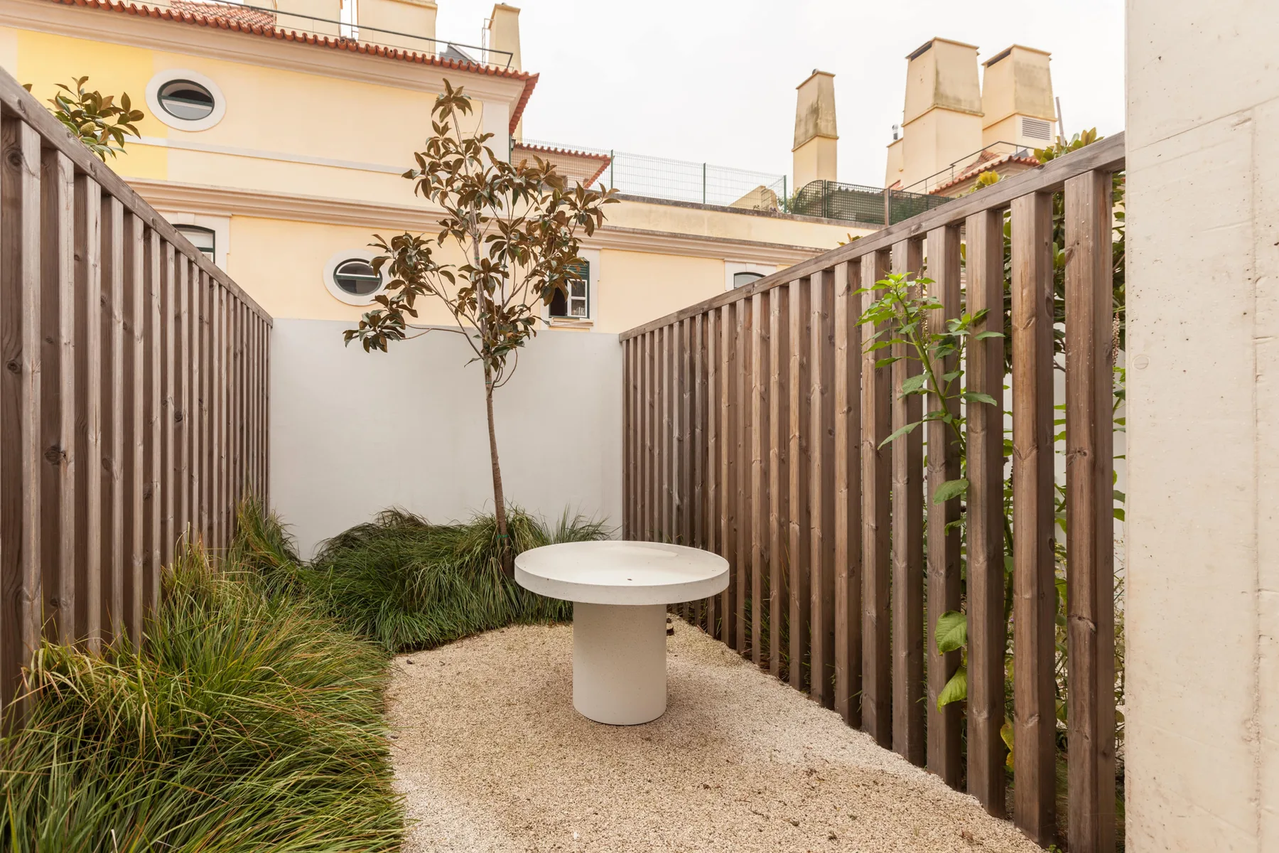 A small outdoor terrace has a round table, plants, and wooden fencing.