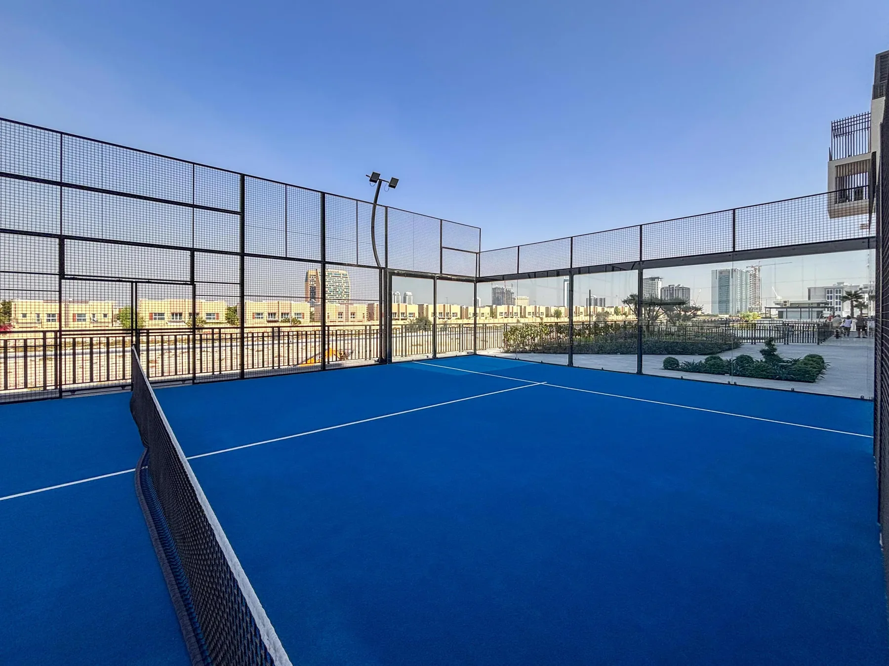Una cancha exterior azul está rodeada de cercas altas, con edificios a lo lejos.