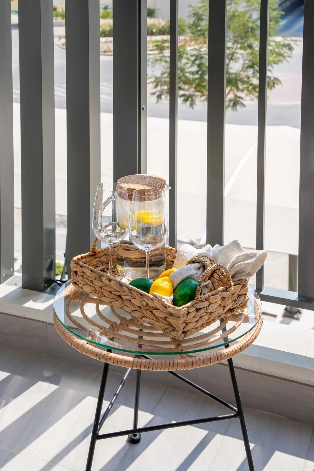 Una mesa pequeña junto a una ventana tiene una cesta de mimbre con fruta y una jarra de vidrio.