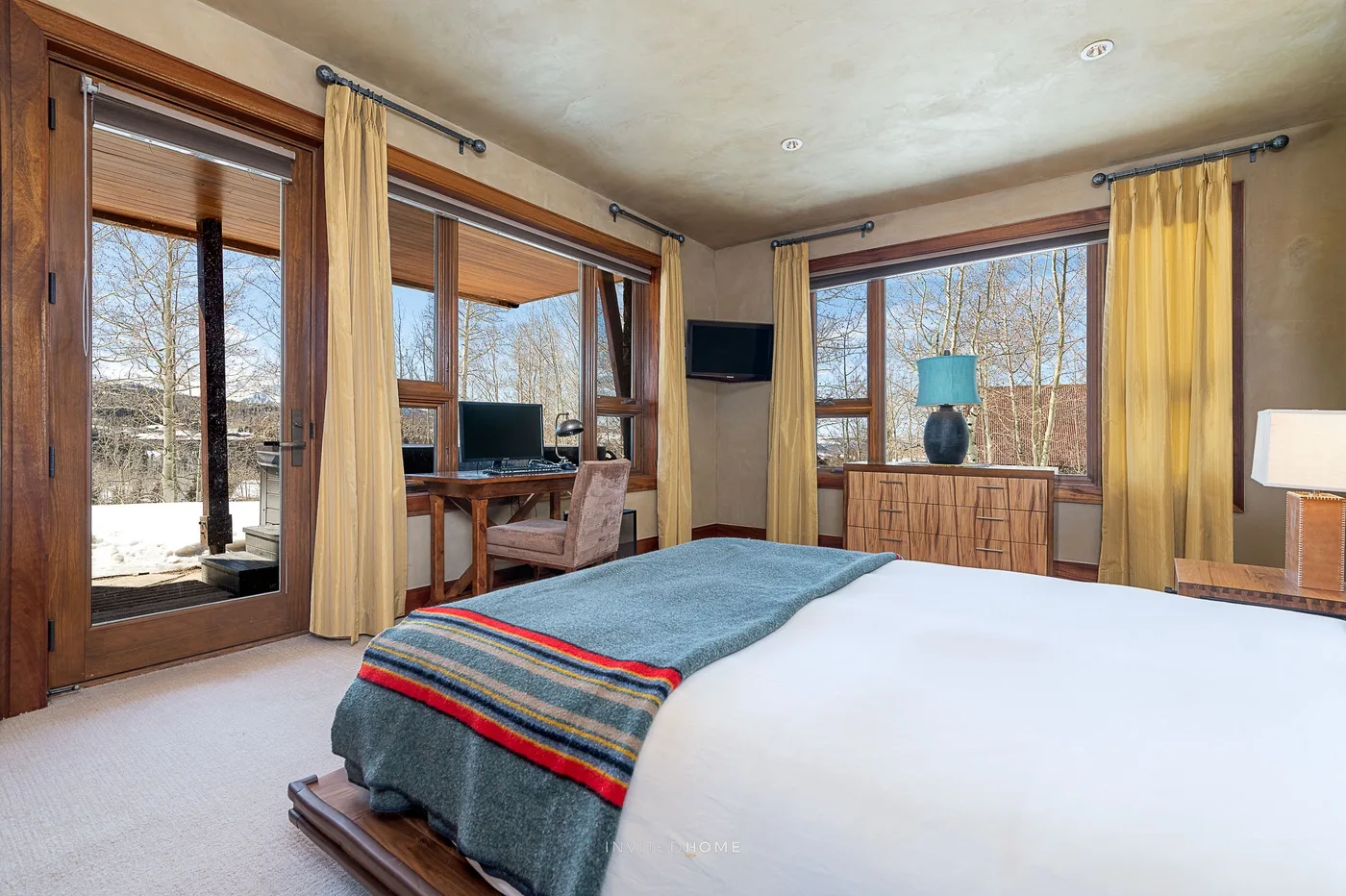 Un dormitorio con una cama, un escritorio y amplias ventanas con vistas a los árboles.