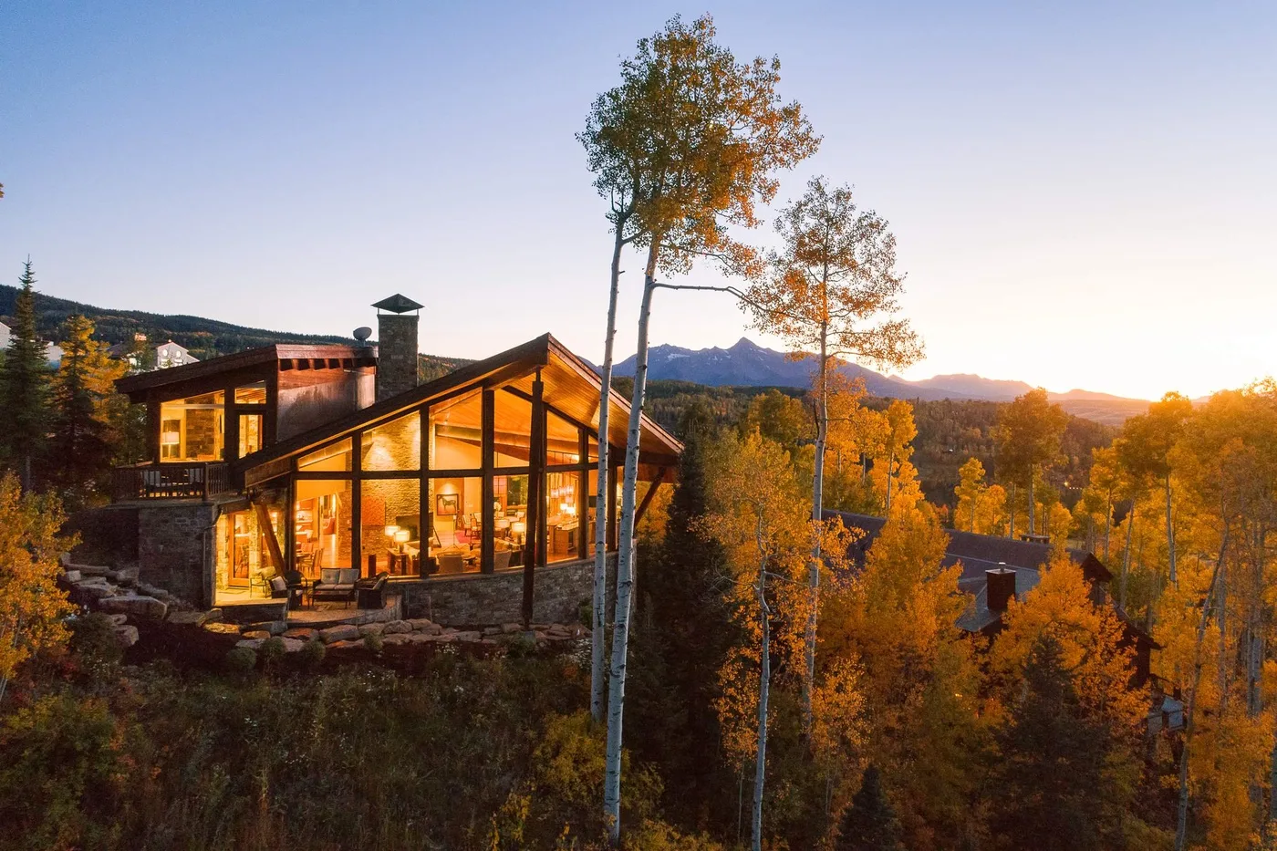 A warmly lit lodge sits among autumn trees with mountains in the distance.