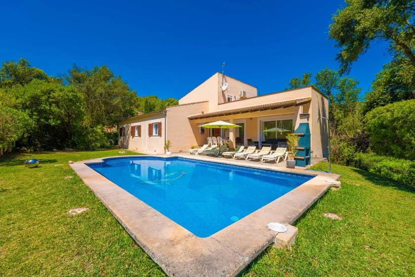 A swimming pool beside a house, surrounded by a green lawn and trees.