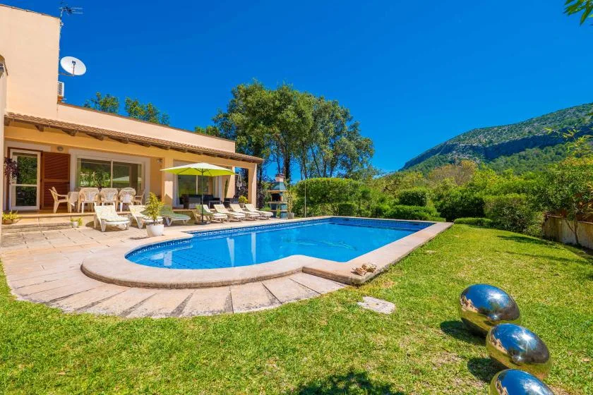A swimming pool and patio sit beside a house, with green hills in the background.