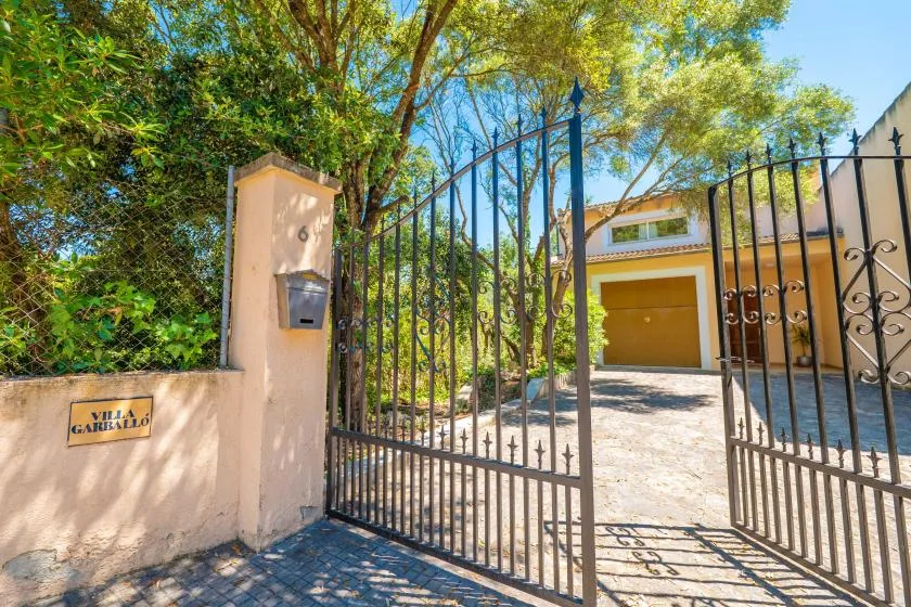 An open metal gate leads to a driveway and garage beneath leafy trees.