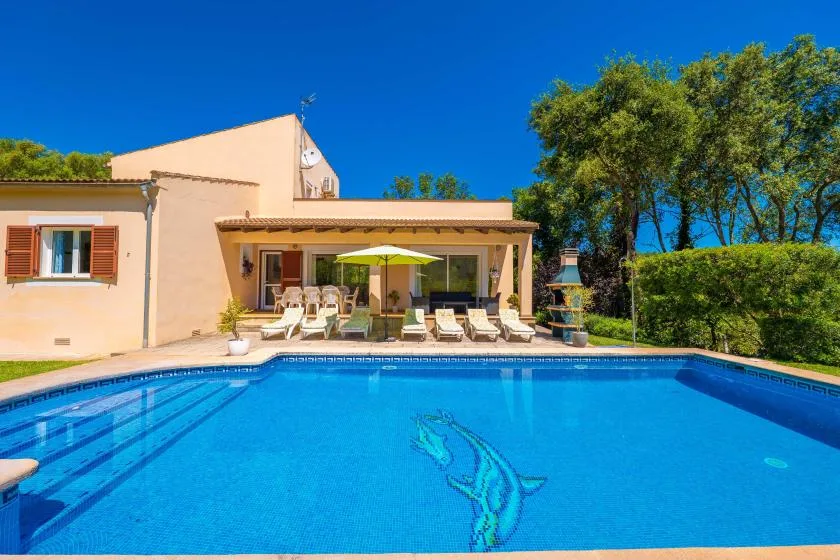 A swimming pool in front of a house, with lounge seating beneath a patio canopy.