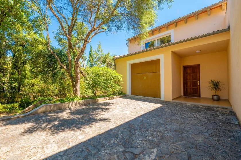 A yellow house with a large garage door and a paved driveway beneath leafy trees.