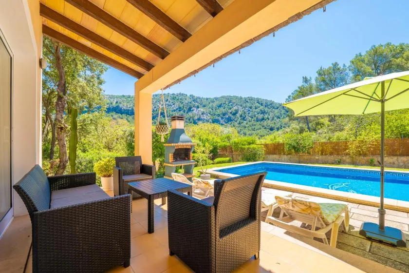 A covered patio with seating overlooks a swimming pool and green hills.