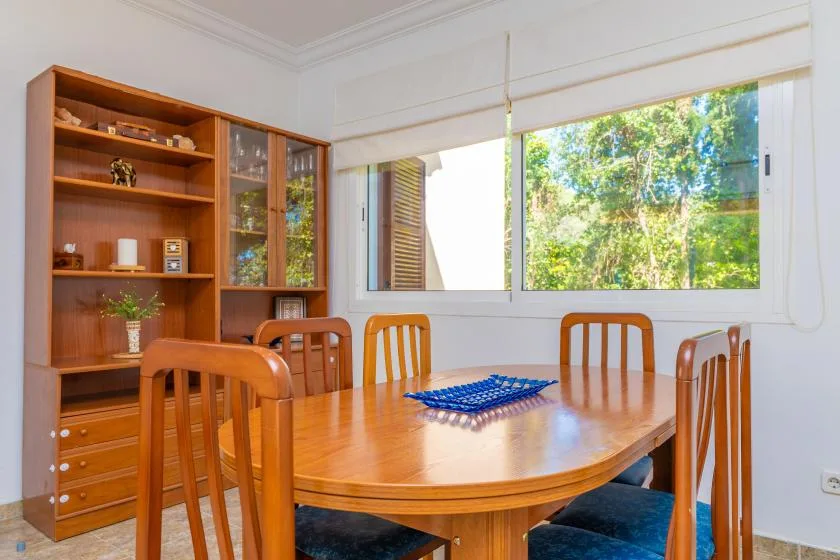 A wooden dining table and chairs sit beside a large window overlooking greenery.